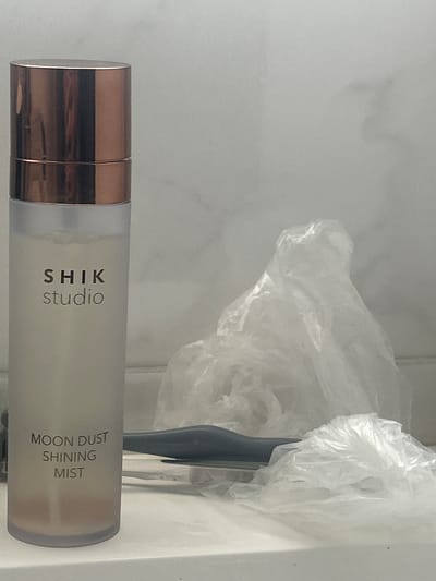 The image shows a bottle of "SHIK studio MOON DUST SHINING MIST" standing on a white surface, possibly a countertop. The bottle is translucent with a rose gold cap and contains a light-colored liquid with some sediment at the bottom. To the right of the bottle are several pieces of crumpled white plastic, appearing to be packaging or discarded materials. The background is a light gray, textured surface, possibly a tiled wall, with some soft lighting. The scene is indoors and appears to be a simple product shot or a casual placement of the item. There is no indication of the city of Нижние Муллы, Russia, or any people or actions occurring in the image.
