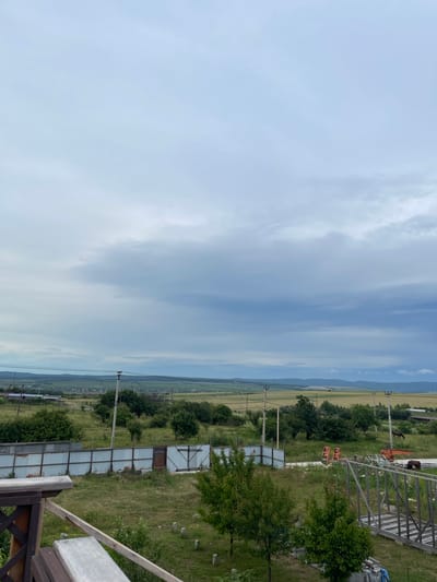 The image depicts an outdoor scene with a cloudy sky and a rural landscape. In the foreground, a wooden structure with railings is visible, possibly a balcony or deck. Beyond this, a grassy area with scattered trees and bushes is present, bordered by a corrugated metal fence with a gate. In the middle ground, the landscape transitions to fields and more trees, with power lines and poles dotting the area. In the distance, rolling hills and a village or town can be seen. There are also some construction-related objects, including an orange crane or similar machinery. A horse is visible grazing in a field in the middle-right portion of the image. The overall impression is that of a somewhat undeveloped, rural area with evidence of both nature and human activity.
