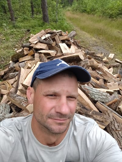 A man wearing a blue baseball cap and a light gray t-shirt takes a selfie in front of a large pile of chopped firewood. He is smiling slightly and looking directly at the camera. The firewood is stacked behind him, with some pieces appearing to be bark-covered logs and others cleanly cut pieces. The background shows a wooded area with green trees and foliage, and a dirt path running through the grass. The scene appears to be in a rural or natural setting, possibly on the outskirts of the Цярэшкавіцкі сельскі Савет in Belarus, suggesting a domestic or outdoor work environment.