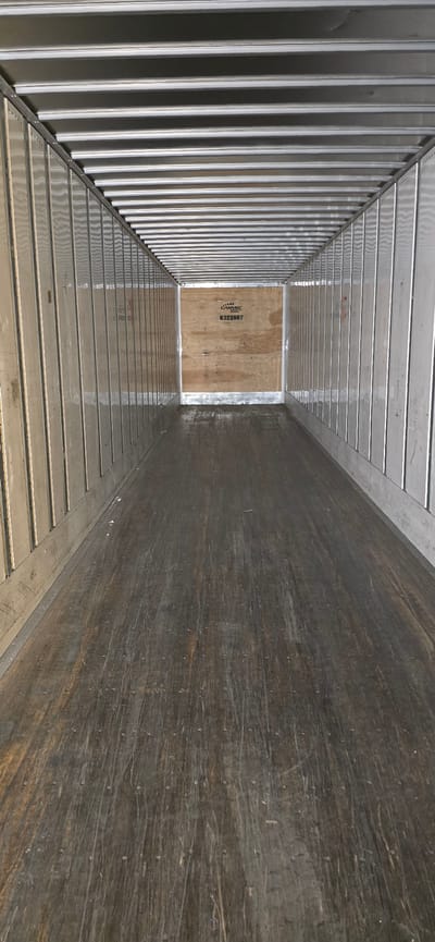 The image shows the interior of an empty truck trailer. The floor is made of wooden planks, showing signs of wear and tear with numerous scuffs and marks. The walls are constructed with vertical corrugated metal panels, and the ceiling features a series of parallel metal beams. At the far end of the trailer, on the back wall, a circular logo with text is visible, along with a number "8322867". The overall impression is one of a utilitarian space, clean but showing signs of previous use, located in Drummondville, Canada.