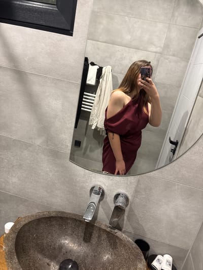 The image depicts a person taking a mirror selfie in a modern bathroom. The bathroom features large grey tiles on the walls and a dark grey countertop. A stone sink with chrome faucets is visible in the foreground. The person is wearing a maroon off-the-shoulder dress and is holding a phone to take the picture. In the reflection, a towel rack with white towels and a black garment is visible. The overall aesthetic suggests a contemporary and minimalist design. This scene is located in Dolno Cherkovishte, Bulgaria.
