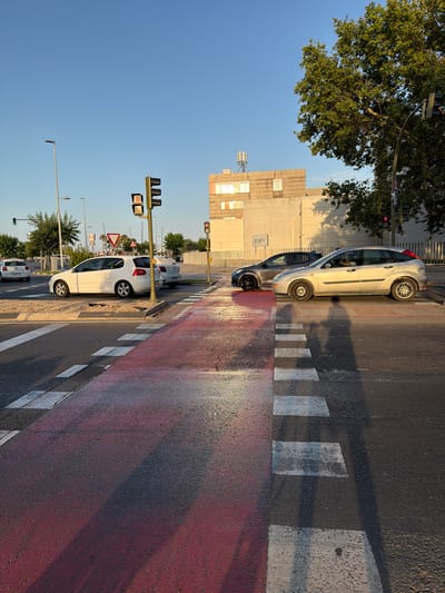 The image depicts a street scene in Castelló de la Plana, Spain, during what appears to be late afternoon or early evening due to the low angle of the sun casting long shadows. The sky is clear and blue, indicating fair weather. The scene is dominated by a pedestrian crossing with a bright red painted surface, bordered by white stripes.

Several cars are visible. In the foreground, a white hatchback is parked, its rear facing the viewer. Behind it, another car, possibly silver, is also parked. Further down the street, a silver sedan is stopped at the pedestrian crossing, with another car partially visible behind it. Traffic lights are present, one showing a red signal, suggesting vehicles are stopped.

The background features a multi-story building with numerous windows and a distinct architectural style, common in urban European settings. Trees line the street, with one large tree prominently featured on the left side of the frame, its branches extending over the road. A cell tower is visible in the distance above the building.

The overall impression is one of a typical urban environment in Spain, with the sun highlighting the wet surface of the road, possibly indicating recent rain. There are no people visibly present in the scene. The image is oriented vertically.