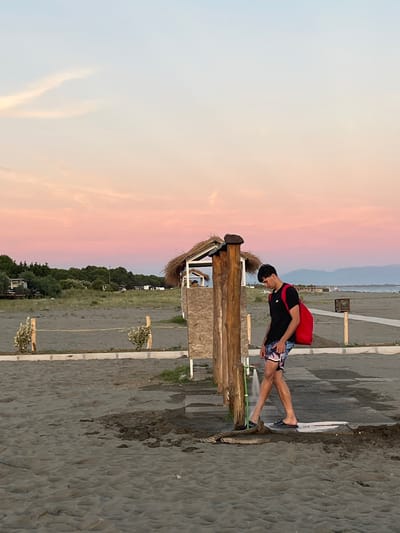 A young man with dark hair is standing on a sandy beach, rinsing his feet under a shower. He is wearing a black t-shirt and patterned shorts, with a bright red backpack slung over his left shoulder. The shower is attached to a wooden post, and water is spraying onto the sandy ground, creating a small puddle. In the background, there is a small, thatched-roof structure that appears to be a changing room or restroom. Further back, a line of trees separates the beach from a low-lying area with some buildings and vehicles. The sky above is a gradient of soft pinks and blues, indicative of a sunset or sunrise. The location is likely near the city of Ulcinj, Montenegro, which is close to Pistula.
