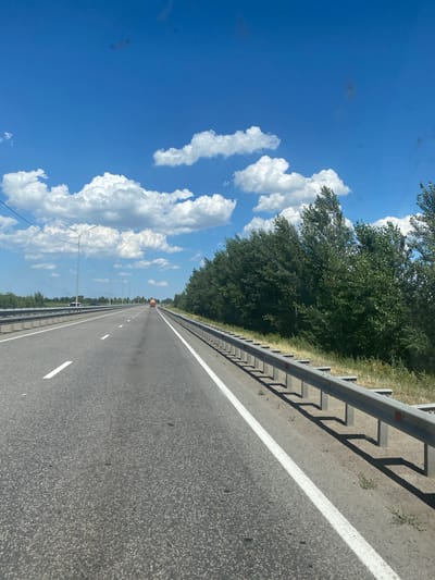 The media file depicts a highway scene on a clear day with a bright blue sky and fluffy white clouds. The road is asphalt, marked with white lines, and has a metal guardrail on the right side. A dense line of green trees borders the highway on the left. In the distance, a few cars and a bright orange vehicle, possibly a construction or maintenance truck, can be seen on the road. Tall lampposts with dangling wires line the side of the highway. The sun appears to be high in the sky, suggesting it is daytime. The overall impression is one of a travel or transit scene. There is no visible text in the image. The location context provided is "Aqsu city administration, Kazakhstan".