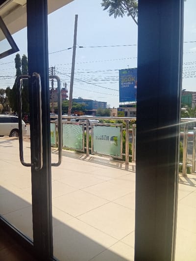 The scene depicts an exterior view observed through a set of dark-framed glass doors equipped with chrome pull handles, from inside a building in Murang'a, Kenya. The foreground shows a light-tiled outdoor patio area directly outside the doors, bordered by a stainless steel railing system featuring frosted glass panels, some displaying reverse text, possibly "natru." Beyond this railing, a partial view of a light-colored vehicle is visible parked on the left. The background reveals an urban environment under a clear, bright blue sky. Overhead utility lines crisscross the frame, accompanied by a tall, slightly leaning utility pole on the left. A prominent blue billboard on the right advertises "GET UPTO 300K IN MOBILE LOANS." Other commercial structures and signs, including "SAYARI" and a sign mentioning "OPTICARE," are discernible in the mid-distance. Strong sunlight illuminates the scene, casting distinct shadows of the railing onto the tiled patio. No individuals are clearly visible or engaged in specific actions.