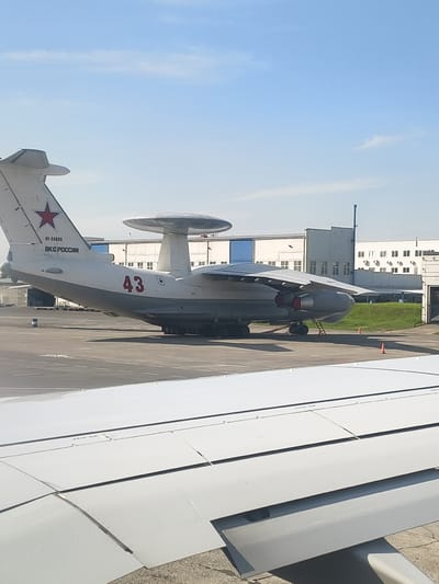 The image shows a large aircraft with a distinctive radar dome on top, parked on a tarmac at an airport in Yemelyanovo, Russia. The aircraft is predominantly white with gray accents. On the tail, a red star and the text "RF-50608" and "ВКС РОССИИ" are visible, indicating it belongs to the Russian Aerospace Forces. The number "43" is prominently displayed in red on the side of the aircraft's fuselage. In the foreground, a portion of an airplane wing, viewed from another aircraft, is visible, suggesting the photo was taken from an airplane window. Several buildings, likely airport structures, are in the background, along with a grassy embankment. The sky is blue with a few wispy clouds. Orange traffic cones are placed on the tarmac.