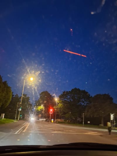 The image shows a night scene in Stafford, United Kingdom. A road with white lane markings stretches into the distance, illuminated by streetlights and car headlights. Traffic lights are visible, with one set showing a red light. Trees line the roadside. The image is taken from inside a vehicle.