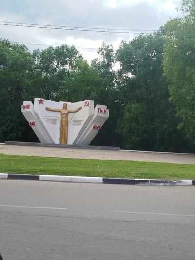 The image shows the "Front to Rear" monument in Noginsk, Russia. The monument consists of a central female figure with outstretched arms, flanked by white triangular structures with the words "Front" and "Rear" written in Russian, along with the years 1941 and 1945. A red star and the hammer and sickle symbol are also visible on the structures. The monument is situated on a grassy area bordered by a black and white curb, with a road in the foreground. Trees form a backdrop to the monument. The sky is overcast.