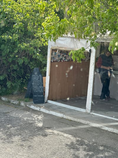 The image depicts a roadside stand in Ταύρη, Greece, partially obscured by foliage. The stand features a white frame and a wooden interior. The back wall is covered with small rectangular items, possibly business cards or similar memorabilia. A chalkboard menu stands outside the stand, listing items such as "ΦΙΛΕΤΑΚΙ" (3.50), "ΣΟΥΒΛΑΚΑΚΙ" (3.50), and "Freddo Espresso" (1.80). A reflection of a person is visible in the glass pane of the stand. The surrounding environment includes lush greenery and a paved roadside.