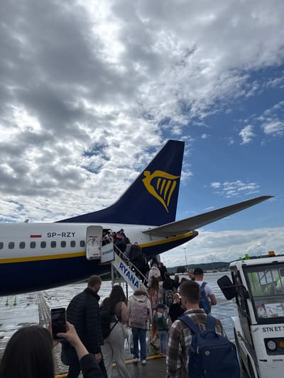 In Balice, Poland, passengers are boarding a Ryanair airplane on the tarmac. The airplane's tail, featuring the distinctive Ryanair logo, is prominently visible against a cloudy sky. A set of stairs leads to the open aircraft door, with a queue of people ascending. Some individuals are holding luggage, while others are taking photographs with their mobile phones. In the foreground, a person is capturing the scene with their phone. A tug vehicle is parked next to the aircraft, its purpose unclear from the image. The ground appears wet, suggesting recent rain. The overall scene depicts the process of boarding an aircraft at an airport.