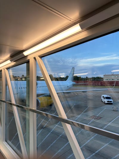 The image depicts an airport jet bridge with an airplane visible through the glass. The jet bridge has a light fixture illuminating the interior. Outside, an airplane with yellow engine nacelles and a distinctive tail design is parked on the tarmac. A white van is also present on the tarmac. The sky is partly cloudy. The scene is likely at Gatwick Airport, which serves the city of Crawley, United Kingdom.