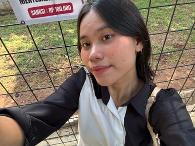 In Cileduk, Indonesia, a young woman with dark hair and light skin takes a selfie outdoors. She is wearing a black and white shirt, and a gold strap from a bag is visible on her shoulder. Behind her, a black metal fence encloses an area with dry grass and dirt. A sign with Indonesian text and a red rectangle stating "SANKSI: RP 100.000" is partially visible in the upper left corner of the image.