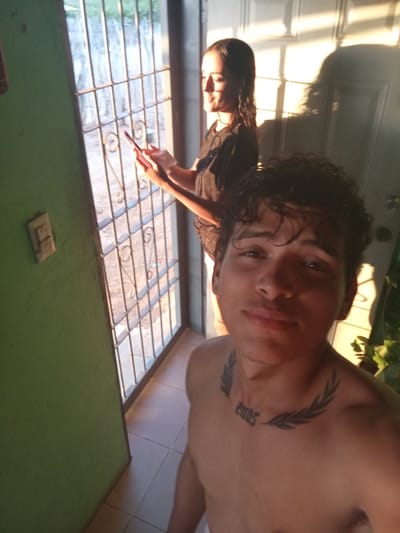 In a sunlit doorway in La Guardia, Venezuela, a young man with curly dark hair and a "2005" tattoo on his chest is taking a selfie. He is shirtless, facing the camera with a slight smile. Behind him, a young woman with dark hair pulled back is standing near a barred window, holding a red object, possibly a phone, and looking to her left. The room appears to be a home with tiled flooring and a green painted wall. Sunlight streams through the window, casting long shadows and illuminating dust motes in the air.
