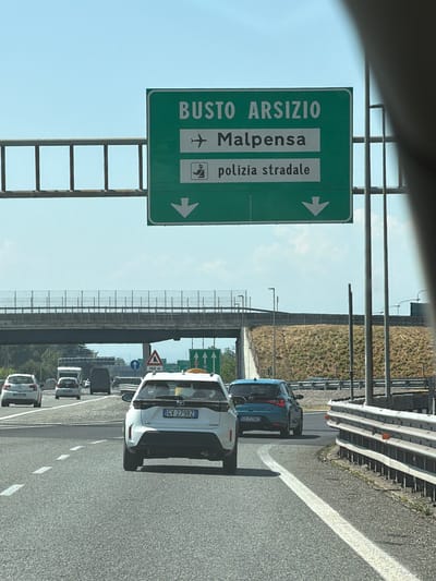 The image depicts a highway scene in Olgiate Olona, Italy, under a clear blue sky. A green directional sign is prominently displayed, indicating "BUSTO ARSIZIO" and "Malpensa" with an airplane icon, suggesting Malpensa Airport is in that direction. Below this, it reads "polizia stradale" (highway police) with a corresponding icon, and two downward-pointing arrows indicate the lanes to take.
In the foreground, a white SUV with the license plate "CV 279RZ" is in the right lane, partially obscuring a blue hatchback with the license plate "CH 722VZ" in the lane to its left. Several other vehicles, including white cars and a van, are visible further ahead on the highway. An overpass with a safety fence runs across the middle ground of the image. The highway is marked with white dashed lines. The surrounding environment features grassy embankments and the metal framework of highway infrastructure.