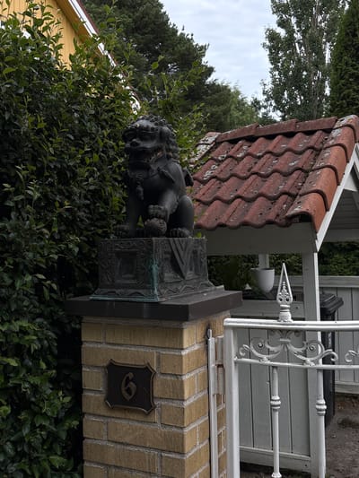 The image shows a decorative statue of a lion, likely a guardian lion, placed on top of a brick pillar. The lion statue is dark and appears to be made of metal, possibly bronze, with intricate detailing. The pillar is constructed from light yellow bricks and features a decorative metal crest with the number "6" prominently displayed.
Behind the pillar, a white ornate fence or railing is visible, suggesting this is an entrance or boundary to a property. The fence is made of wrought iron with decorative scrollwork. A portion of a red-tiled roof with weathered edges is also present, possibly belonging to a nearby building or structure. Green foliage, including dense bushes and trees, surrounds the statue and pillar, indicating an outdoor setting. A yellow house can be seen in the background on the left side of the image.
The image appears to be taken during the daytime, under an overcast sky, as indicated by the soft, diffused lighting and the appearance of the sky. There is no specific activity or event taking place; the scene depicts a static view of the property's decorative elements. The overall impression is one of an established, perhaps slightly older, residential or public property in a green, leafy environment. There is no visible text other than the number "6" on the crest.