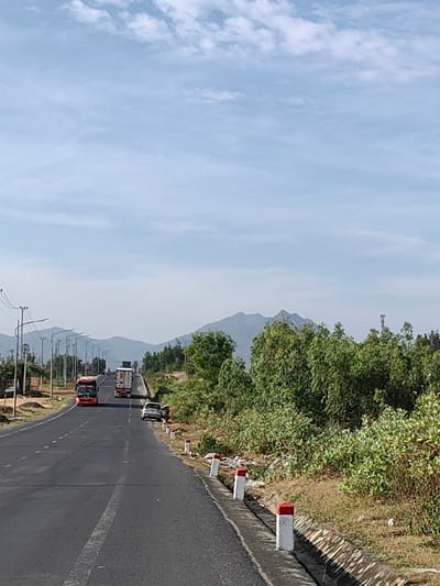 This image captures a multi-lane asphalt highway in Tuy Phong 4, Vietnam, under a bright, partly cloudy sky. The road, featuring at least two lanes for traffic moving away from the viewer, is marked by a dashed white line and extends towards a mountainous horizon. On the right shoulder, a white sedan is stationary, positioned partially off the pavement and into the grassy verge next to a concrete drainage ditch. This ditch is bordered by white and red-topped road markers. Further down the road, a red passenger bus occupies the left lane, followed closely by a white semi-trailer truck. Another large truck is visible much further in the distance. The left side of the road is lined with utility poles supporting power lines and streetlights. Both sides of the highway are flanked by dense green trees and various shrubs. Hazy, rugged mountains form the backdrop of the scene. No individuals are visible.