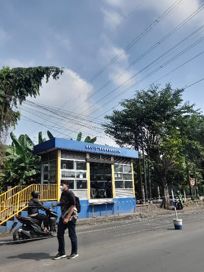 The scene is on a road in the Kecamatan Waru, Indonesia. A man wearing a mask walks towards the camera on a paved road. He is carrying a blue backpack and is in the foreground. A small blue and yellow structure is to the left of the man. The structure has a sign on the roof that says "KALI-BUNGGASIH". It appears to be a security checkpoint, with windows that have security bars. There are several people inside the structure. A motorcycle with a person on it is parked behind the man on the right side of the road. A blue traffic cone is to the right of the motorcycle. A small tree is behind the motorcycle. Numerous power lines crisscross the sky. The sky is clear and blue, with white clouds. There is green vegetation to the left of the man. There is a yellow railing in front of the vegetation.