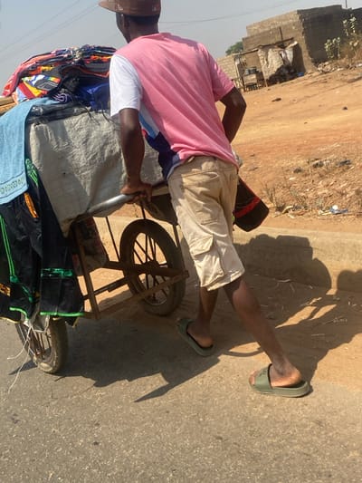 An individual, appearing male, is observed pushing a two-wheeled utility cart laden with assorted textiles and garments along an unpaved road in Lapai, Nigeria. The subject wears a brown baseball cap, a pink and white short-sleeved t-shirt, beige cargo shorts, and dark green slide sandals. A gold-colored bracelet is visible on the subject's right wrist. The cart, constructed with a metal frame, carries folded clothing items of various colors and is partially covered by a light gray tarp; a blue towel and black garments with green trim hang from its left side. The ground transitions from a dark asphalt surface in the immediate foreground to reddish-brown, dry earth. Strong sunlight from the upper right casts prominent shadows of the subject and cart. The background reveals sparse, dry vegetation and several utilitarian structures constructed from concrete blocks and corrugated metal, indicating a non-urbanized setting.
