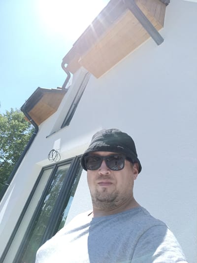 A man in a grey t-shirt and black bucket hat with sunglasses stands in front of a white building, possibly under construction or recently renovated, with wooden eaves and black gutters. He is looking directly at the camera. The building has large windows with dark frames. Green trees are visible to the left of the frame. The sky is bright blue with harsh sunlight creating lens flare in the upper portion of the image. The overall scene is brightly lit, suggesting it is daytime. The location is identified as Rataje, Czechia, but no specific street or landmark is visible in the image to confirm this.