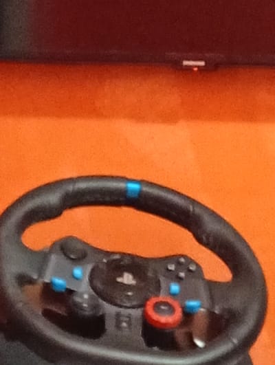 The image shows a black PlayStation racing wheel with a blue central marker on the wheel rim and various buttons, including a red one. The wheel is positioned in front of an orange background, with the lower edge of a black rectangular object (likely a TV) visible at the top. The environment appears to be indoors. There is no indication of the location being Malko Chochoveni, Bulgaria.