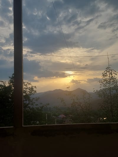 The image depicts a sunset over a mountainous landscape in Areni, Armenia. The sky is filled with dramatic clouds, tinged with the golden hues of the setting sun, which is partially obscured by the clouds and appears low on the horizon. In the foreground, a dark, unfinished concrete structure frames the view, with a vertical wooden post on the left side. To the right, silhouetted trees with dense foliage add depth to the scene. In the middle ground, rolling hills are covered with greenery, and hints of houses and structures suggest a village or small town nestled within the valley. The overall atmosphere is serene and natural, characteristic of the Armenian countryside. There are no people or animals visible in the frame. The image is captured from a slightly elevated viewpoint, looking out over the expansive scenery.