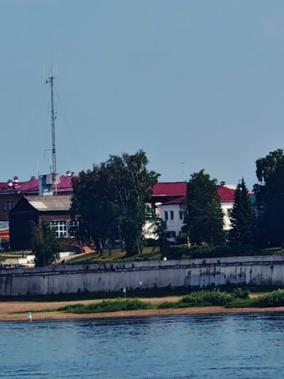 The image shows a view of the Yenisei River in Yeniseysk, Russia. On the riverbank, there is a white concrete embankment, followed by a grassy area and a beach. A single person wearing a blue shirt and white shorts is standing on the beach. Beyond the embankment, several buildings are visible, including a dark-colored building with white-paned windows and a larger white building with a red roof. A tall radio tower stands near the dark-colored building. The sky is clear and pale blue. The overall impression is of a sunny day in a Russian city by the river.