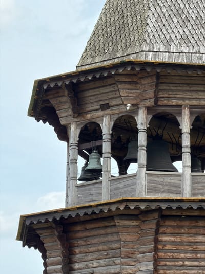 The image shows a close-up view of a wooden bell tower in Nevskii Parkleskhoz, Russia. The bell tower is constructed from logs and has a multi-tiered roof covered in wooden shingles. Multiple bells are visible within the open arches of the tower. A small white security camera is mounted on one of the wooden beams. The sky is overcast.