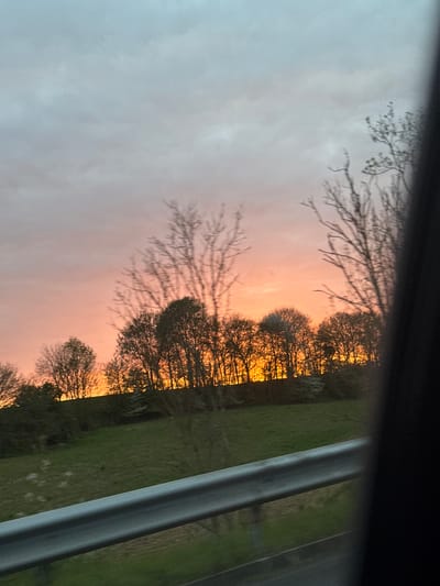 The image shows a view through a window, likely from inside a vehicle or building. The visible scene consists of a sunset sky with an orange and pink hue peeking through overcast gray clouds. Silhouetted trees are situated against the sunset. Beyond the trees, there's a green field or grassy area. A portion of the window frame is visible on the right side of the image. Without additional context, I cannot verify whether this image was taken in Saint-Quentin, France.