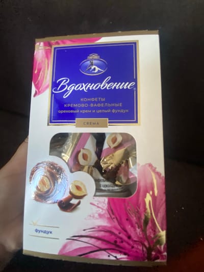 This is a product image of a box of chocolates. The box is predominantly white with blue and pink floral designs and gold accents. The brand name "Вдохновение" (Inspiration) is written in elegant script on a blue background. Below this, it states "Конфеты кремово-вафельные с ореховым кремом и целым фундуком" which translates to "Cream-waffle candies with nut cream and whole hazelnuts." There is a smaller label that reads "CREMA". The box has a window revealing several individually wrapped chocolates, some of which show a hazelnut and chocolate coating. There is also an image of a hazelnut with a chocolate swirl. On the lower left, the word "фундук" (hazelnut) is written. The background is dark and out of focus, appearing to be a textured surface, possibly a couch or blanket. The lighting suggests an indoor environment, likely a home. The image does not contain any information indicating its location in the city of Akhtyrskiy, Russia, nor does it depict any people or actions.