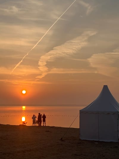 At sunset on the beach in Surakovo, Russia, the sky is painted in warm hues of orange and yellow, with wispy contrails streaking across the clouds. The sun, a bright orb, casts a golden reflection on the calm water. In the foreground, a white tent stands on the sand. A group of silhouetted figures are gathered near the water's edge, some appearing to be taking photos. One figure is seated in a foldable chair. The scene is tranquil, capturing a moment of shared enjoyment of the natural beauty.