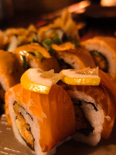 A close-up shot of sushi rolls, possibly part of a meal, is presented. The rolls are predominantly salmon, characterized by their vibrant orange color and glistening texture. Each roll is meticulously prepared, featuring white rice encased in seaweed and topped with thin slices of lemon. The lemon slices add a bright yellow accent and a hint of freshness to the dish. Some rolls appear to have a creamy sauce drizzled over them, adding another layer of visual appeal. The background is dark and out of focus, suggesting an indoor setting, perhaps a restaurant or a home during an evening meal, given the lighting. The composition focuses on the appetizing appearance of the sushi, highlighting its colors and textures. There is no visible text in the image. The overall impression is one of a delicious and carefully crafted culinary experience.