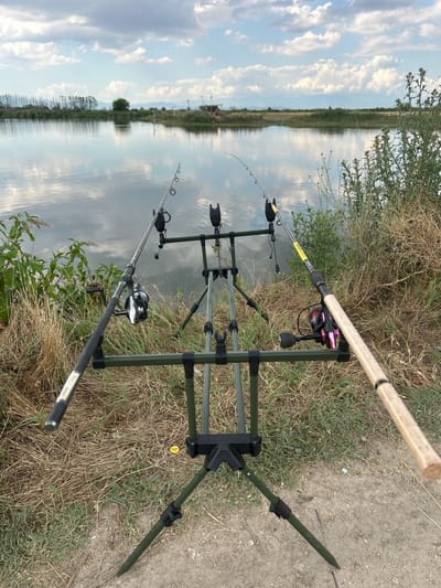 In the city of Vodyano, Bulgaria, fishing rods are set up on a tripod stand beside a calm body of water, reflecting the sky and clouds. The scene is framed by dry grass and some green vegetation. The time of day appears to be late afternoon or early evening, given the lighting.