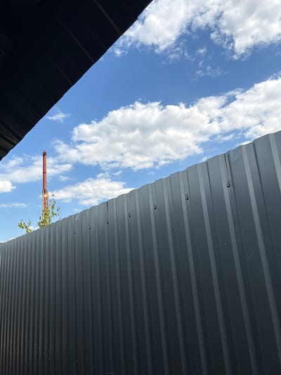 The image is a low-angle shot looking up at a clear blue sky with scattered white clouds. Two sections of dark, corrugated metal roofing are visible, angled towards each other from the bottom left and top right of the frame. The roofing appears to be part of an outdoor structure, possibly a shed or lean-to, providing shade.

In the gap between the two roofing sections, a red, cylindrical object, resembling a chimney or a pipe, is visible against the sky. Some green foliage, likely from a vine or small tree, is growing around the base of this object. The lighting suggests it is daytime, and the clear sky indicates fair weather. The scene appears to be an outdoor setting, possibly in a yard or industrial area. There are no people visible in the image, nor any visible text.