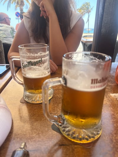 The image depicts a vibrant outdoor scene from a cafe or restaurant, likely in a sunny, coastal location such as Puerto Del Carmen, Spain. The view is from a seated perspective, looking across a table towards the sea.
In the foreground, two large, handled beer mugs rest on a light brown, possibly wooden, table surface. The upper mug, closer to the camera and partially consumed, clearly displays the "Mahou" brand name on its label, featuring a substantial head of foam. Below it, a fuller mug is visible, branded "San Miguel" with "CERVEZAS SINCE 1890" printed beneath, also topped with a foamy head. Both beers are light amber lagers.
Two individuals are partially visible. On the left side, the arm and part of the upper body of a person with long, dark hair and a white top are seen, resting on the table. Further back and to the left, the profile of a bald man wearing glasses and a light-colored, patterned short-sleeve shirt (reminiscent of a Hawaiian shirt) is visible, looking towards the background.
The background is dominated by clear blue skies and bright sunlight. Several lush palm trees are prominent, suggesting a tropical or subtropical environment. Beyond the trees, a sparkling blue expanse of ocean or sea is visible. A red and white circular road sign, possibly a "no entry" or "stop" sign, can be seen amidst the greenery. The bright, clear weather and setting evoke a relaxed holiday atmosphere, suggesting daytime enjoyment of drinks by the sea.