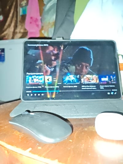 The image depicts a close-up view of a tablet device displaying a video player interface with movie thumbnails and playback controls. The tablet is positioned on a wooden surface, likely a table, in front of a blurred background containing items of clothing. To the left of the tablet is a black computer mouse, and to the right is a white object, possibly another peripheral. The tablet screen shows the movie "Summertime (film, 2001)" at the top, and below it, a section labeled "Recommendations" with thumbnails for various films. The overall scene suggests a casual setting, likely indoors, where someone is watching or browsing for movies on a digital device. The context of Akhmetovskaya, Russia, is not visually evident in the provided image.