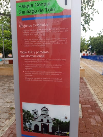 The image is a close-up shot of a signpost with information about Parque Central Santiago de Tolú in Tolú, Colombia. The sign has a green top section with the text "Parque Central Santiago de Tolú" in white letters. Below this is a red section with white text detailing the history of the park. The first section of text describes "Orígenes Coloniales" (Colonial Origins) and mentions the founding of Tolú in 1535 by Alonso de Heredia, its role as a center for religious celebrations, and its adherence to the model of Spanish colonial cities. The second section, "Siglo XIX y primeras transformaciones" (19th Century and first transformations), details changes made during the republican era, including the establishment of gardens and paths for a more organized urban character, and the park becoming a symbol of local identity surrounded by administrative and religious buildings. A small photograph is attached at the bottom of the red section, showing a white church with red accents and bell towers, with palm trees visible behind it. The signpost is a light gray metal structure. The background shows a paved area with brickwork, some trees, and the edge of a blue railing and walkway. The lighting suggests an overcast day.