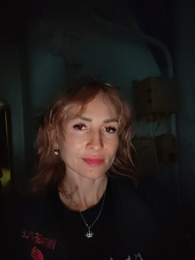 A woman with shoulder-length reddish-blonde hair and light skin is looking at the camera. She is wearing a black shirt with a silver crown-shaped pendant necklace and a diamond stud earring in her visible left ear. The background is dark and appears to be an interior space with some industrial elements, such as conduits and boxes on the wall. The lighting is dim. The location is not discernible from the image.