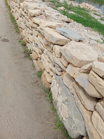 A low, rough-hewn stone wall, constructed from irregularly shaped, light-colored rocks, runs along the edge of a dirt path. The wall is not meticulously built, with stones of varying sizes and thicknesses stacked loosely, some appearing to be in disarray. Small green weeds and dry grass sprout from between the stones and along the base of the wall. The dirt path is a muted brown, showing some slight discoloration and a few scattered dried plant stems. In the background, beyond the wall, is a slope covered with sand, pebbles, and more green vegetation. Further in the distance, a body of water is visible, with its surface reflecting the ambient light. This scene is located in Sol-Iletsk, Russia.