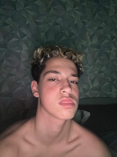 The image displays a head-and-shoulders portrait of a young male, likely in his late teens or early twenties, positioned against a dark, textured background. The subject has short, curly hair with prominent blonde or light brown highlights, styled away from his forehead. His eyes are dark brown, and he is looking directly at the camera. His lips are distinctly pursed. His complexion is fair, with a reddish tint visible on his cheeks. He appears to be shirtless, revealing his clavicles and upper chest; a small, dark mole or blemish is visible on his right collarbone.

The background is a dark teal or dark green wall featuring a repetitive, three-dimensional geometric pattern composed of triangular or faceted shapes, suggestive of decorative paneling or wallpaper. The lighting is dim, appearing to originate from the front or above the subject, casting subtle shadows and highlighting parts of his hair and shoulders. The setting is an indoor location within the city of Strazhitsa, Bulgaria. No other individuals or specific objects are identifiable in the immediate vicinity.