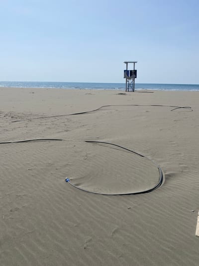 An outdoor scene captures a wide, unoccupied sandy beach in Ulcinj, Montenegro, under a clear, bright blue sky. In the immediate foreground and extending into the midground, a thick, dark-colored flexible hose or cable is laid across the rippled sand, forming a prominent curve and extending linearly towards the background. The end of the hose closest to the viewer is fitted with a blue and white cylindrical connector. The sand exhibits distinct wind-formed ripples and undulations. In the midground, a light-colored wooden lifeguard tower, featuring a blue back panel and a shaded roof structure, stands vertically on the sand, positioned towards the water's edge. The ocean, appearing calm with a visible horizon line, forms the background. No individuals or specific actions are depicted within the frame.