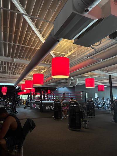 The image shows the interior of a gym, likely located in Enschede, Netherlands, identified by the visible website address "WWW.BIGGYM.NL" on a banner. The gym features an industrial-style ceiling with exposed pipes and beams, illuminated by large, cylindrical red pendant lights. In the foreground, a man in a black tank top and blue shorts is seated on a weight machine, facing away from the camera, with a dark towel draped over the seat. To his left, another person is visible on a different weight machine. The middle ground of the gym contains a boxing ring with red ropes and a black banner. Various exercise machines, including cardio equipment like treadmills and elliptical trainers, are arranged throughout the space. Other gym-goers are seen in the background, some using equipment and others walking. The overall environment is well-lit and appears to be a functional fitness facility.