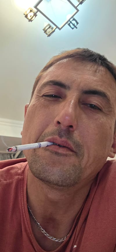A close-up, head-and-shoulders shot, captured from a low angle, shows a Caucasian male with light brown hair and stubble. He is wearing a reddish-orange t-shirt and a silver chain necklace. The man is holding a lit cigarette horizontally in his mouth, with smoke gently rising from it. His expression is neutral, with his eyes partially closed and wrinkles visible around them. The background consists of a white ceiling with a modern light fixture. There are no identifiable landmarks or contextual elements in the image that would suggest a specific location within the city of Gori, Georgia.