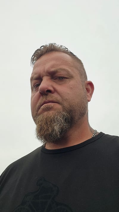 A low-angle, close-up, outdoor portrait shows a Caucasian male with a stern expression, looking down and to the left. He has short, dark hair that is wet and swept back, a thick beard with grey streaks, and a shaved head on the sides. He is wearing a black t-shirt with a dark graphic print visible on the chest. A silver chain necklace is visible around his neck. The background is a pale, overcast sky. The image was taken in Milton, Canada.