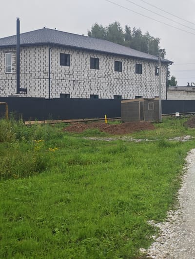 The image shows a partially constructed building with a grey shingle roof and white cinder block walls in Vorsma, Russia. The building has several black-framed windows and a black chimney pipe on the left side. A dark grey fence runs along the front of the property. In front of the fence, there is a small, dark grey shed with a wooden door and a small window. The area in front of the shed and building is uncultivated, with piles of dirt and overgrown green grass and weeds. A gravel path is visible on the right side of the image. The sky is overcast.