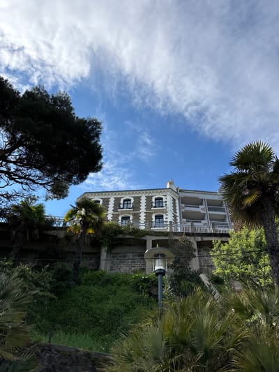 The image shows a building with a white facade and a stone foundation located at the coordinates 48.634, -2.05, placing it in the city of Dinard, France. The building, built in the style of a large house, is located on a steep hillside covered in vegetation. There is a stone wall below the building, with a small lamp post protruding from it. The building is obscured from view in parts by a large, leafy, dark green tree on the left side of the image. A palm tree sits to the right of the building, while another leafy green tree occupies the lower right corner of the image. There is a blue sky with many clouds in the background.