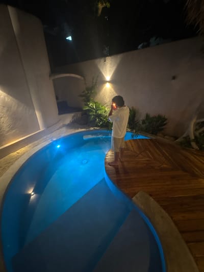 This nighttime image depicts an outdoor private pool area in El Nido, Philippines. A single individual, an adult male with dark hair, stands barefoot on a circular wooden deck at the pool's edge. He wears a white short-sleeved shirt and light-colored shorts. The male is positioned with his back mostly to the camera, his head slightly turned to the left, and is actively lighting an unseen object held between both hands near his face, indicated by a bright orange flame.
The irregularly shaped pool contains blue-lit water, with at least one visible underwater light source on the left side. Light-colored coping borders the pool, and steps are visible descending into the water on the left. Surrounding the pool are white, textured stucco walls, illuminated by warm-toned, upward and downward-facing wall lights, particularly prominent on the left. Tropical vegetation, including palms and various leafy plants, is strategically placed around the pool area and against the walls. In the upper left background, an arched alcove is visible within the wall structure. The wooden deck features planks arranged in a distinct pattern, curving with the pool's edge. The sky above is dark and indistinct.