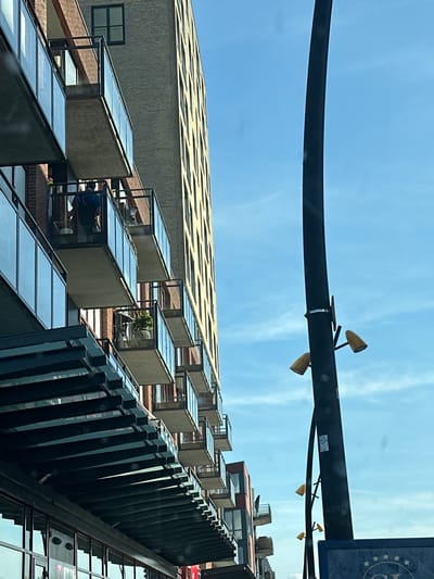 This is a vertical, eye-level, outdoor shot of a modern brick and glass building with balconies, situated in Heerhugowaard, Netherlands. The building's facade features repeating balconies with glass railings. On one of the balconies, a person wearing a dark shirt and backpack is visible. The sky is bright blue with some wispy clouds. To the right of the building, a tall, black lamppost with two yellow light fixtures is prominent. The bottom of the image shows a dark awning structure extending from the building.
