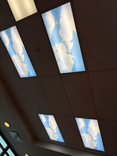 The image shows a drop ceiling with several illuminated panels depicting a blue sky with white, fluffy clouds. One panel is a plain, bright light. The ceiling tiles are a muted, brownish-gray color. A light fixture and air vent are visible along the right edge of the frame, near a window. The overall impression is that the room is dimly lit, with the illuminated panels providing the primary light source.