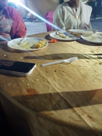 A table draped with golden cloth holds remnants of a meal. Discarded paper plates with food scraps sit beside a black smartphone and a white plastic spoon. Two people are partially visible at the table. One wears an orange shawl and a patterned shirt. The other is dressed in a white outfit and a large golden pendant necklace. A pink chair is located behind the person in white.
