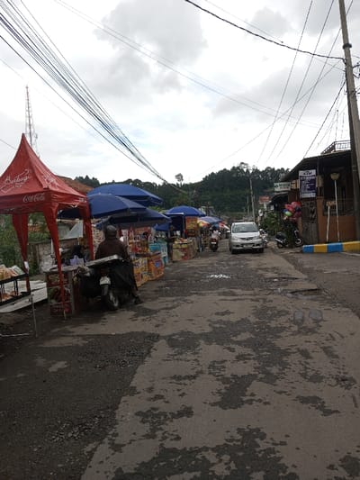 This image depicts a bustling street market scene in the city of Melakudikadu, India, under an overcast sky. The foreground shows a worn asphalt road with numerous cracks and potholes.
On the left, a series of street vendor stalls line the roadside. The leftmost stall is covered by a red canopy tent featuring white lettering and "Coca-Cola" branding. Adjacent to it are multiple stalls sheltered by dark blue umbrellas, displaying various packaged food items and beverages. A person wearing a dark jacket and helmet is seated on a parked motorcycle in front of one of these stalls, facing away from the viewer. Further in the background on the left, a tall communication tower is visible.
The right side of the road features several buildings. One building has a prominent "ATM" sign, while another adjacent structure displays a "Pharmacy" sign with additional text. A utility pole stands prominently, supporting a dense network of overhead electrical and communication wires that crisscross the upper portion of the frame. A cluster of colorful balloons is visible near the pharmacy building. The curb along this side of the road is painted in alternating blue and yellow segments.
In the mid-ground, a silver multi-purpose vehicle (MPV) is parked facing the camera. Another motorcycle is observed further down the road, moving away from the viewer. The background features lush green hills covered with dense foliage, indicating a rural or semi-urban setting with natural elevation. Several individuals are present among the market stalls, engaging in unidentifiable transactions or activities typical of a market environment.